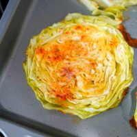 A close-up view of baked cabbage steaks roasted in an oven. The light green slices have crispy, browned edges and are sprinkled with spices, resting on a metallic baking tray.