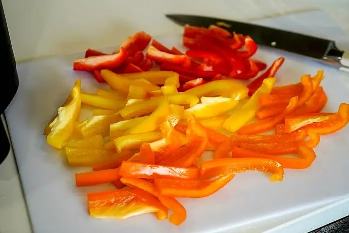 Sliced red, yellow, and orange bell peppers are arranged on a white cutting board, ready for the next step in learning how to blanch bell peppers. A knife is placed nearby on the board.