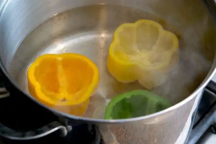 Slices of yellow, orange, and green bell peppers are boiling in a pot of water on the stove, demonstrating how to blanch bell peppers perfectly. Steam rises from the pot.