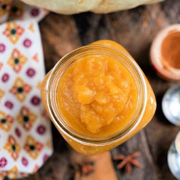 Top view of a jar filled with smooth, orange pumpkin butter made in a pressure cooker on a wooden surface. A patterned cloth is partially visible on the left, with small bowls and star anise in the background.