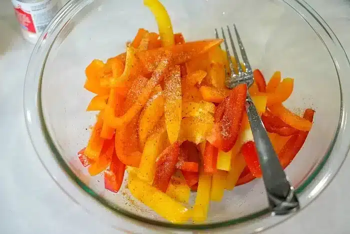 A glass bowl brimming with vibrant, sliced red, yellow, and orange bell peppers is artfully seasoned. If you're curious about prepping them further, learning how to blanch bell peppers can enhance their texture. A fork and spoon are nestled inside the bowl for easy serving.