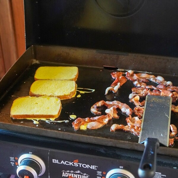 A Blackstone 22 griddle sizzles with bacon strips and bread slices being toasted, nestled next to a spatula.