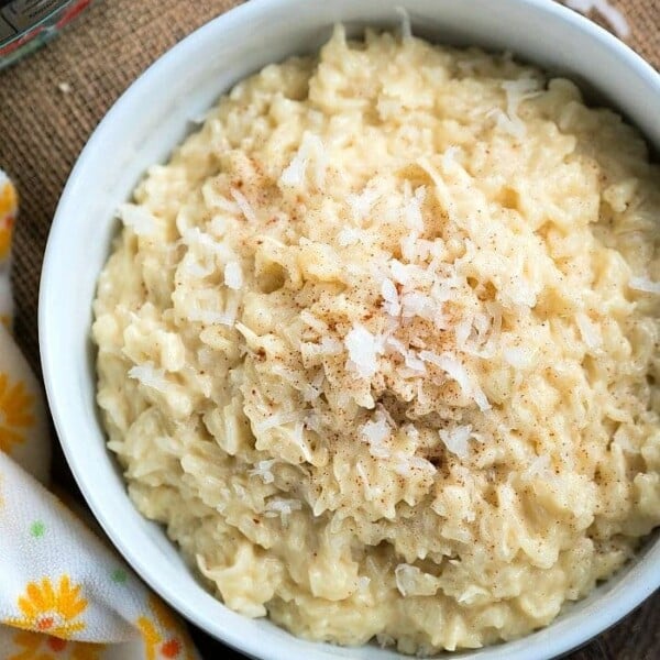 A white bowl filled with creamy pressure cooker coconut rice pudding, topped with a sprinkle of cinnamon and shredded coconut, sits on a burlap surface. A yellow floral-patterned cloth is partially visible beside the bowl.