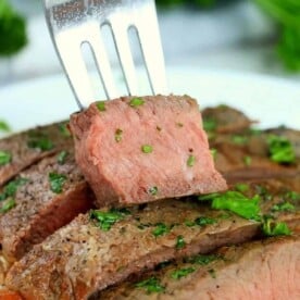 A fork holds a slice of medium-rare steak garnished with chopped herbs. Several slices, cooked from frozen in an air fryer, are arranged on a plate in the background, also sprinkled with herbs, against a backdrop of blurred green foliage.