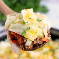 A close-up of a spoonful of unstuffed cabbage roll casserole reveals layers of cooked cabbage, ground meat, diced tomatoes, and melted cheese. Fresh herbs garnish the top. In the background, more casserole awaits in the pan.