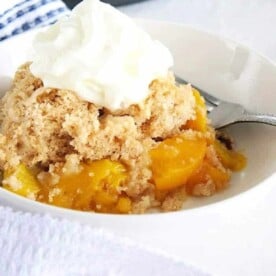 A bowl of instant pot peach cobbler crowned with whipped cream, with a fork waiting eagerly by the side.