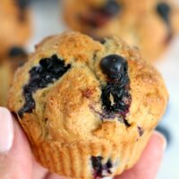 A hand holds a freshly baked blueberry muffin, made effortlessly in an air fryer, with more muffins tantalizingly blurred in the background.