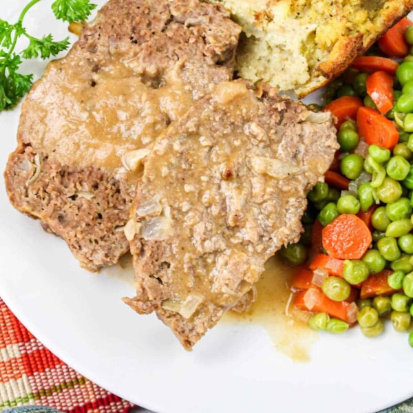 A quick meatloaf recipe served elegantly: a plate with sliced meatloaf, topped with rich gravy, accompanied by vibrant peas and carrots, and a slice of bread on a pristine white dish.