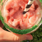 A person enjoys a pink frozen drink adorned with green sugar and sipped through a black and white striped straw. Standing on the grass, sandals peeking from beneath their jeans, they contemplate how to make a peppermint frappuccino for their next treat.