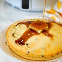 An air fryer pancake made with Bisquick is topped with a pat of butter as syrup is drizzled over it. The golden delight rests on a white plate, with a floral-patterned cloth partially visible in the background, adding a charming touch to this breakfast scene.