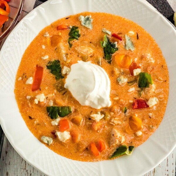 A white bowl brimming with creamy instant pot buffalo chicken soup is garnished with a dollop of sour cream. This savory delight includes chunks of vegetables and herbs, vibrant red and yellow peppers, and bits of blue cheese scattered on top.