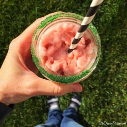 A person enjoys a glass of pink slushy with a black and white striped straw, its rim dusted in green sugar. As they stand on the grass in socks and blue jeans, you might wonder how to make a peppermint frappuccino for an equally delightful treat.