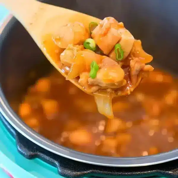 A wooden spoon lifts succulent Pressure Cooker Hoisin Chicken pieces coated in sauce and green onions, hovering above a teal pressure cooker brimming with the same savory dish. This aromatic stew or curry is rich with vibrant onions and fresh herbs.