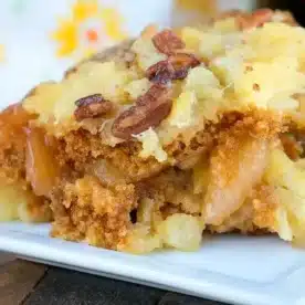 A close-up of a serving of baked apple dessert on a white plate. The gingerbread dump cake features a crumbly topping with pecans and pieces of apple, creating a golden-brown texture. A floral-patterned napkin is partially visible in the background.