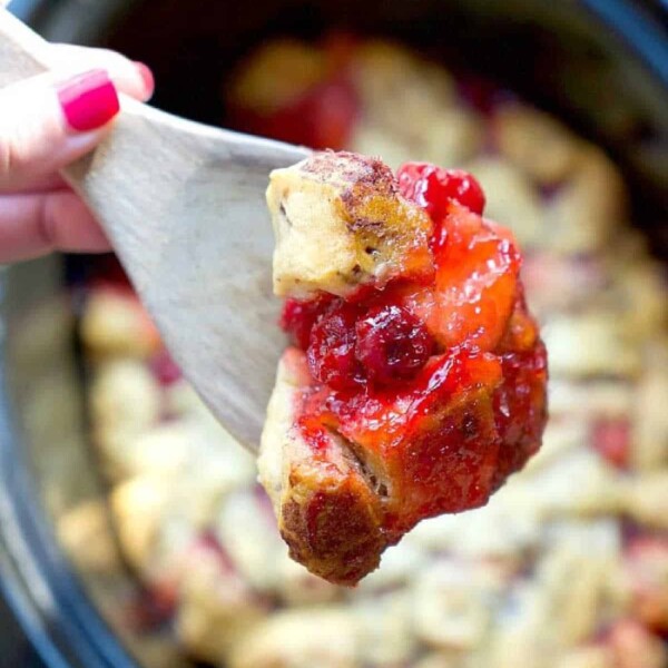 A hand with red nail polish holds a wooden spoon, showcasing a piece of baked dessert resembling cinnamon roll monkey bread, featuring a golden-brown crust and vibrant red fruit filling. In the background, a crockpot brims with more of this delightful treat.