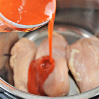 Pouring spicy buffalo sauce over uncooked chicken breasts in a stainless steel pot for a savory Instant Pot feast.