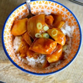 A colorful bowl of crockpot orange chicken topped with sliced green onions, with a fork inside.