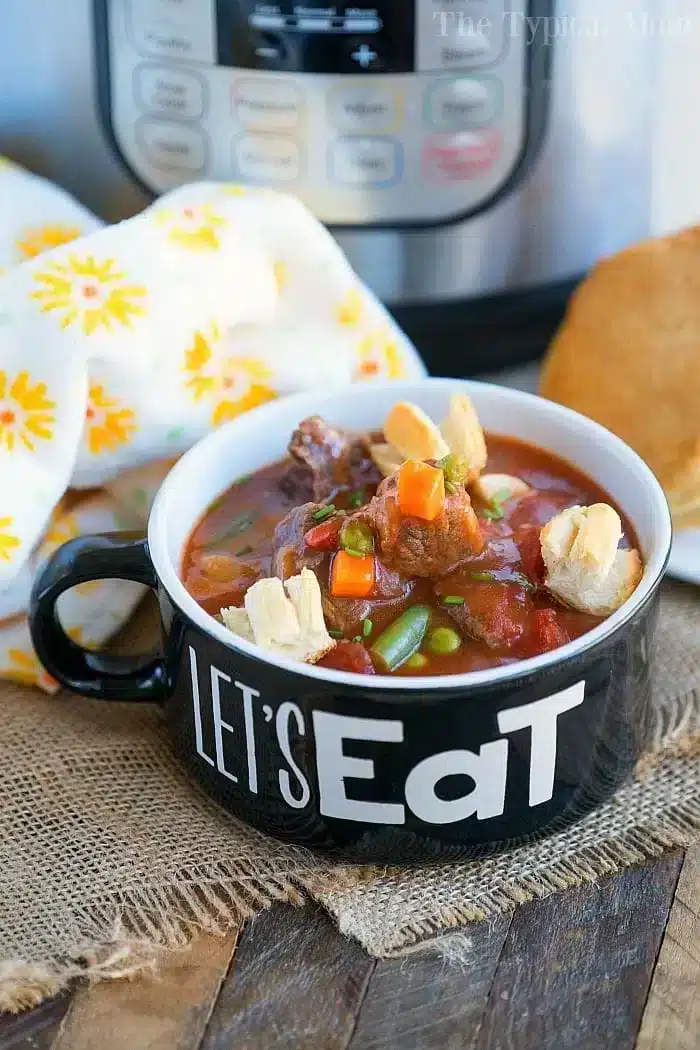 A black mug emblazoned with "Let's Eat" brims with pressure cooker beef pot pie soup, garnished with tiny pie crust shapes. It sits on a wooden surface alongside a white floral cloth, while a pressure cooker lingers in the background.