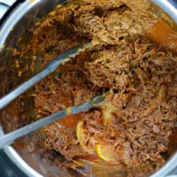 Shredded barbacoa beef in an Instant Pot, with metal tongs on the side.