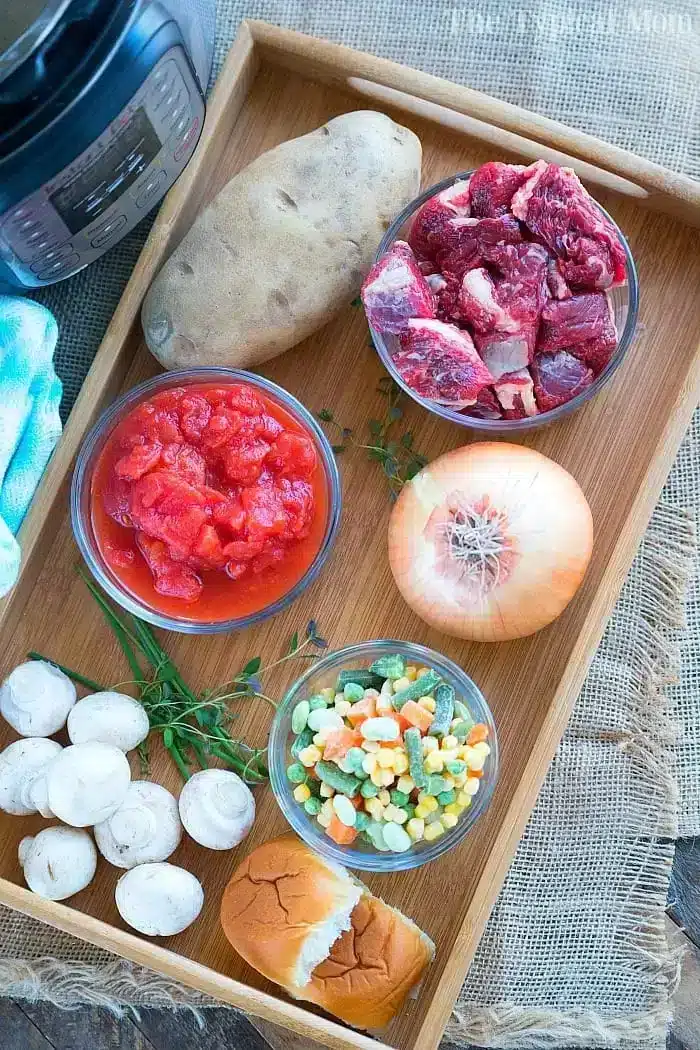 A wooden tray holds the makings of a hearty beef pot pie soup: a whole potato, raw beef chunks, diced tomatoes in a bowl, a whole onion, sliced mushrooms, mixed frozen vegetables, fresh thyme, and a bread roll. An electric pressure cooker is partially visible in the corner.