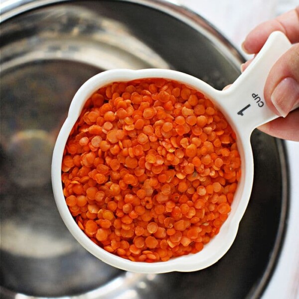 A hand holds a 1-cup measuring cup brimming with vibrant red lentils over a metal bowl, ready for the instant pot.