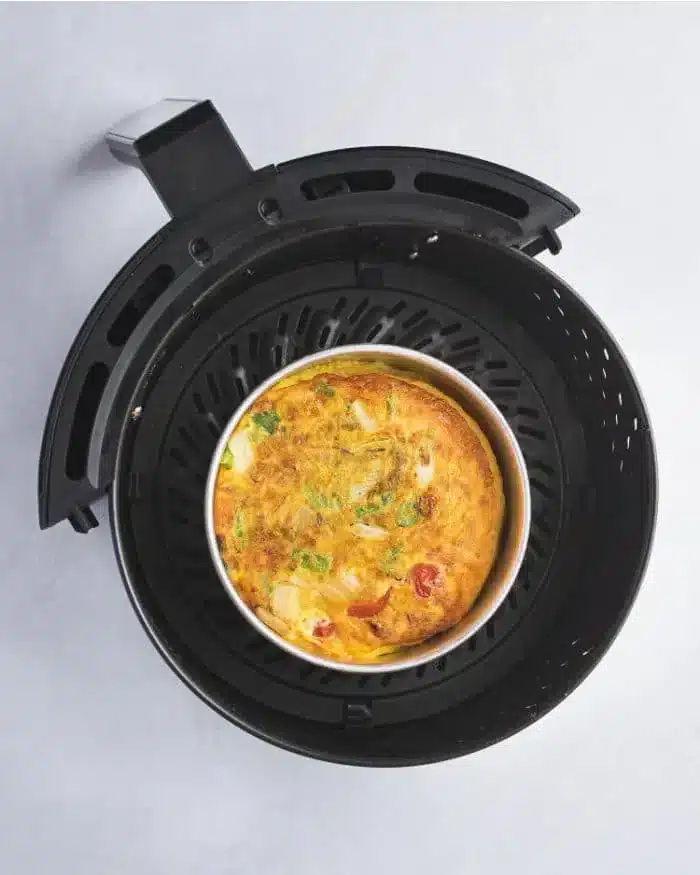 An air fryer with a removable basket contains a delicious quiche, featuring visible ingredients like tomatoes, green herbs, and cheese. The appliance is placed on a light-colored surface.