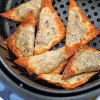 A batch of golden-brown air fryer won tons is neatly arranged in the basket. Their crispy, triangular shapes boast varying shades of deliciousness against the dark surface, highlighting their perfect fry.