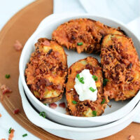 A white bowl contains four air fryer potato skins with crispy cheese and bacon bits, garnished with chopped green onions. One potato skin has a dollop of sour cream. The bowl is placed on a brown placemat with scattered bacon bits around.