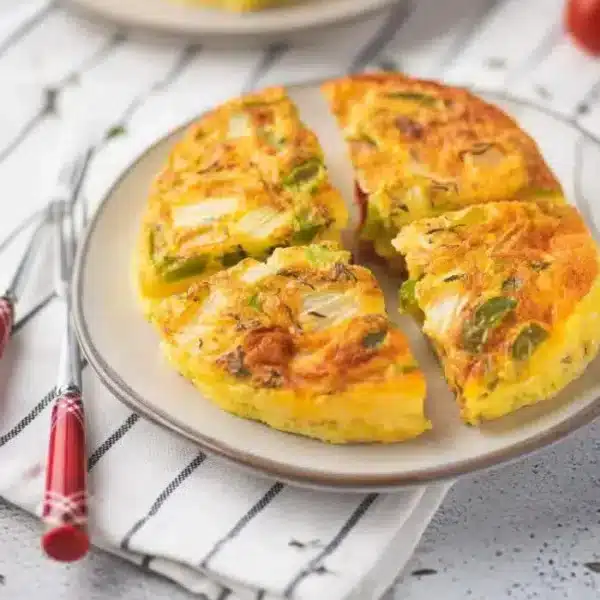 A plate featuring a sliced Ninja Foodi frittata is shown, showcasing visible pieces of onion and green vegetables. Red-handled forks are placed beside the plate, and the background is adorned with a striped cloth.