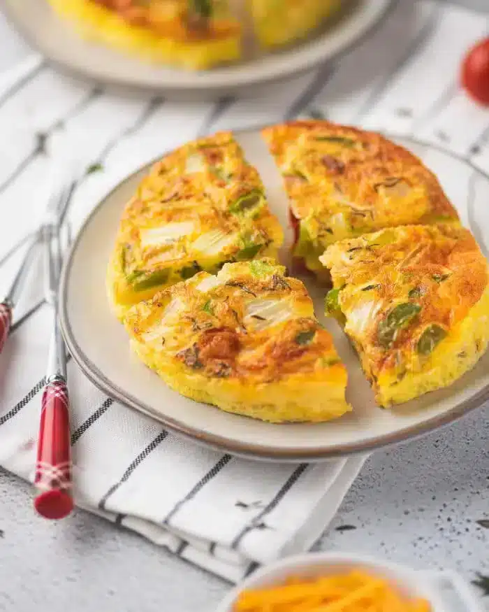 A plate with a sliced air fryer quiche featuring vegetables like onions and bell peppers. The quiche is served on a light-colored plate on a striped cloth with red-handled forks nearby.