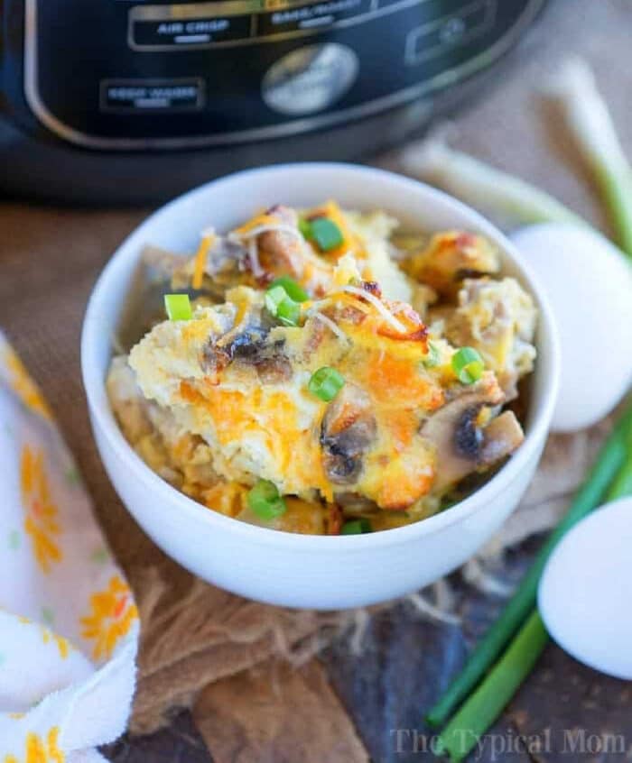 Bowl of cheesy breakfast casserole with green onions, made in the Ninja Foodi, next to eggs and a kitchen appliance in the background.