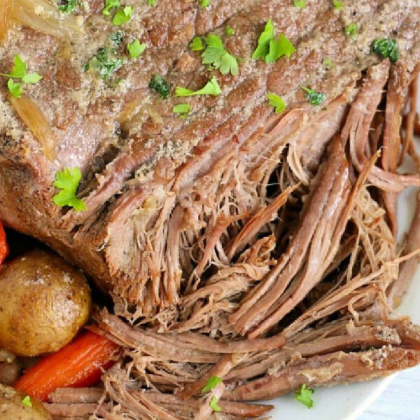 A close-up of a tender Ninja Foodi Roast Beef on a plate. The beef is perfectly cooked, with shredded sections and garnished with fresh parsley. It sits alongside roasted carrots and potatoes. A fork is partially visible at the bottom right corner.