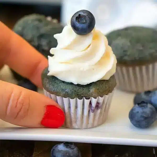 A hand with red nail polish gracefully holds a blue cupcake topped with creamy white frosting and a single blueberry, reminiscent of a bite-sized blueberry pie. More blueberries and similar cupcakes rest invitingly on the wooden surface in the background.