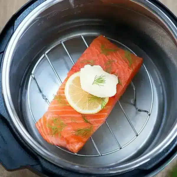 A piece of raw salmon topped with a lemon slice, fresh dill, and butter is placed on a metal rack inside an Instant Pot, showcasing one of the finest pressure cooker fish recipes ready for cooking.