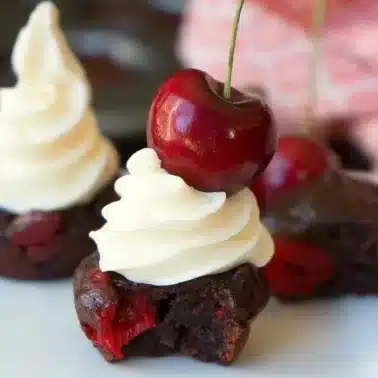 A chocolate brownie bite topped with a swirl of white frosting and a whole cherry. A partial bite reveals a surprise cherry filling, reminiscent of biting into that first scoop of blueberry pie filling. Two similar brownie bites are blurred in the background.