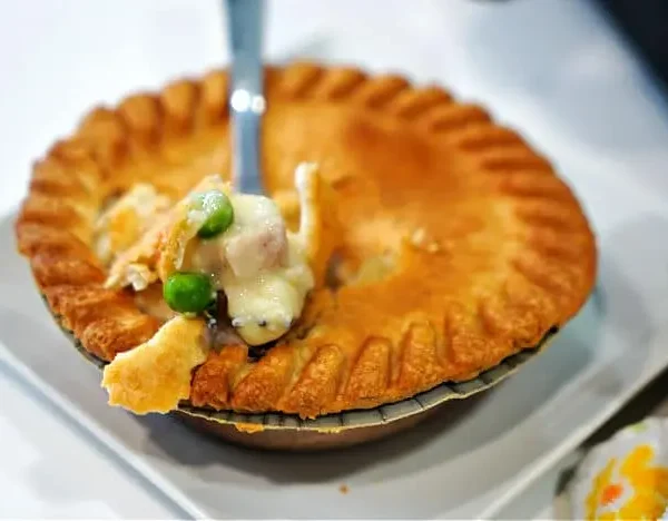 A fork lifts a bite of creamy chicken pot pie with peas and carrots, fresh from a frozen pot pie cooked to perfection in an air fryer. The flaky, golden-brown crust has a beautiful fluted edge, and a patterned cloth is partially visible beside the white plate.