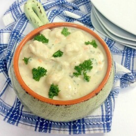 A ceramic bowl filled with creamy instant pot gnocchi soup garnished with parsley sits on a blue and white checkered cloth. A stack of white plates rests in the background.