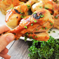 A close-up of a hand holding a grilled chicken drumstick garnished with parsley reveals juicy goodness, reminiscent of flavors unlocked from frozen chicken legs when seamlessly cooked in an instant pot. More drumsticks grace a plate in the background, surrounded by fresh parsley on the wooden surface.