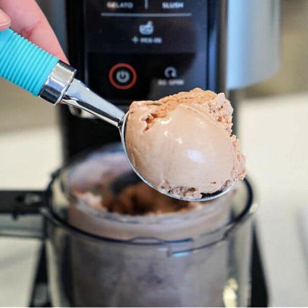 Close-up of a hand holding a blue-handled ice cream scoop with a scoop of chocolate ice cream in front of a Ninja Creami ice cream machine.