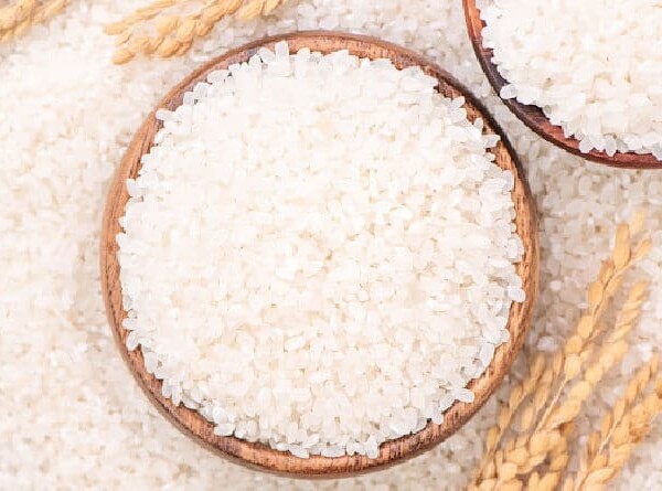 A wooden bowl brimming with long grain white rice rests on a surface sprinkled with rice grains, surrounded by wheat stalks.