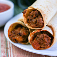 Three beef tacos on a plate with a bowl of salsa in the background, evoking the same delightful crunch as perfectly cooked air fryer flautas.