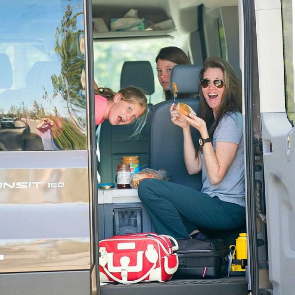 Three people in a van, one savoring a sandwich, another peeking from a seat. A travel bag rests on the floor, filled with essentials for discovering the best vacation spots for kids.