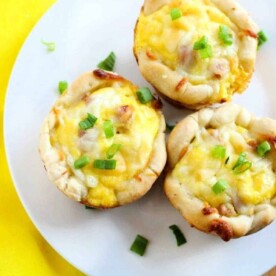 Three mini egg and cheese muffins topped with green onions grace a white plate, evoking the savory delight of ham and cheese biscuits, with a yellow napkin placed nearby.