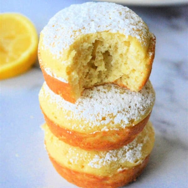 Three lemon Bisquick muffins stacked, powdered sugar dusted on top, with a bite taken from the top muffin. A fresh lemon slice lingers in the background.