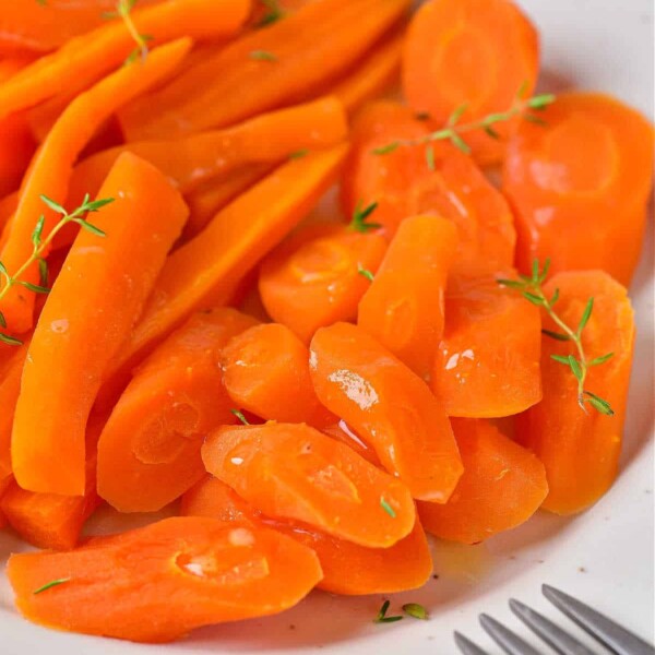 Sliced and glazed carrots with herbs are elegantly arranged on a white plate beside a fork, offering a perfect example of how to cook carrots to enhance their natural sweetness.