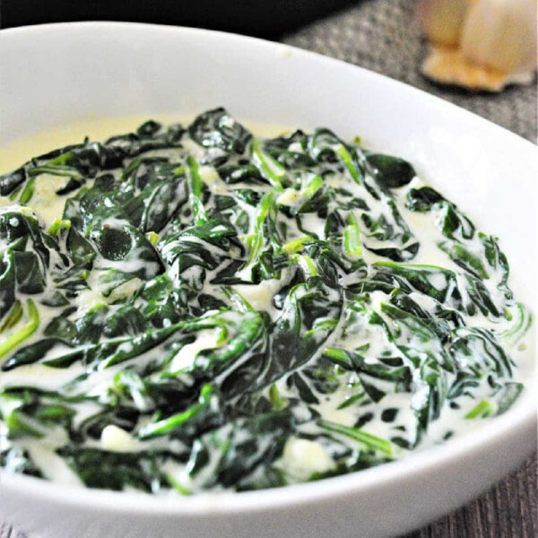 A bowl of simple creamed spinach with heavy cream sits on a wooden table, with garlic in the background adding a rustic touch.