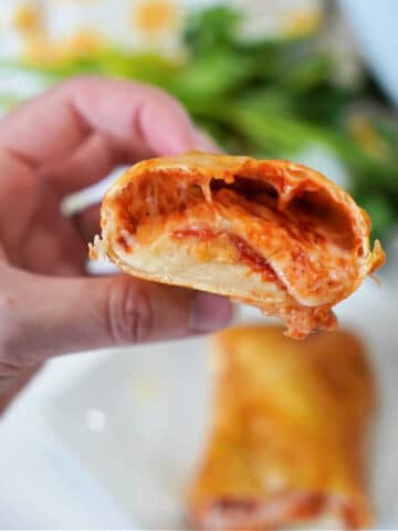 A hand is holding a halved, air fryer hot pocket filled with melted cheese and tomato sauce, showcasing its gooey interior. Another whole roll is visible on a plate in the blurred background, with some green leaves partly visible.