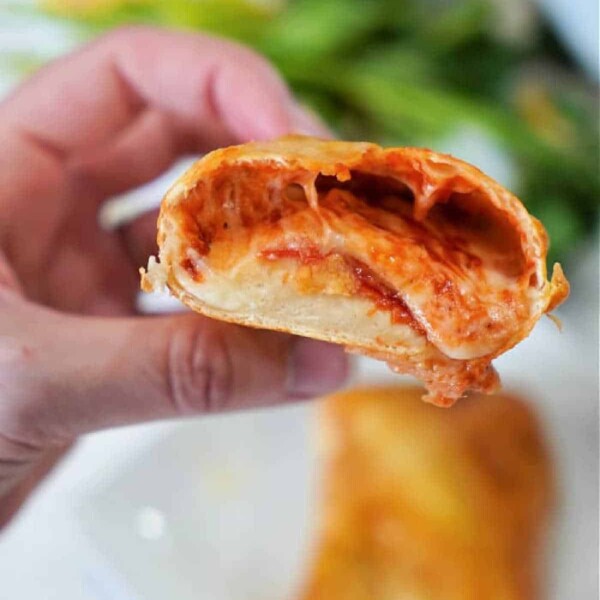 A hand is holding a halved, air fryer hot pocket filled with melted cheese and tomato sauce, showcasing its gooey interior. Another whole roll is visible on a plate in the blurred background, with some green leaves partly visible.