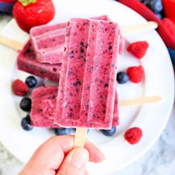 Hand holding a berry popsicle over a plate with more berry popsicles and fresh berries, showcasing the art of how to make homemade popsicles with fresh ingredients.