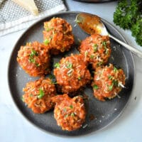 A plate with seven instant pot porcupine meatballs covered in tomato sauce and garnished with chopped herbs. A spoon rests on the plate, while a piece of cheese and parsley linger in the background.